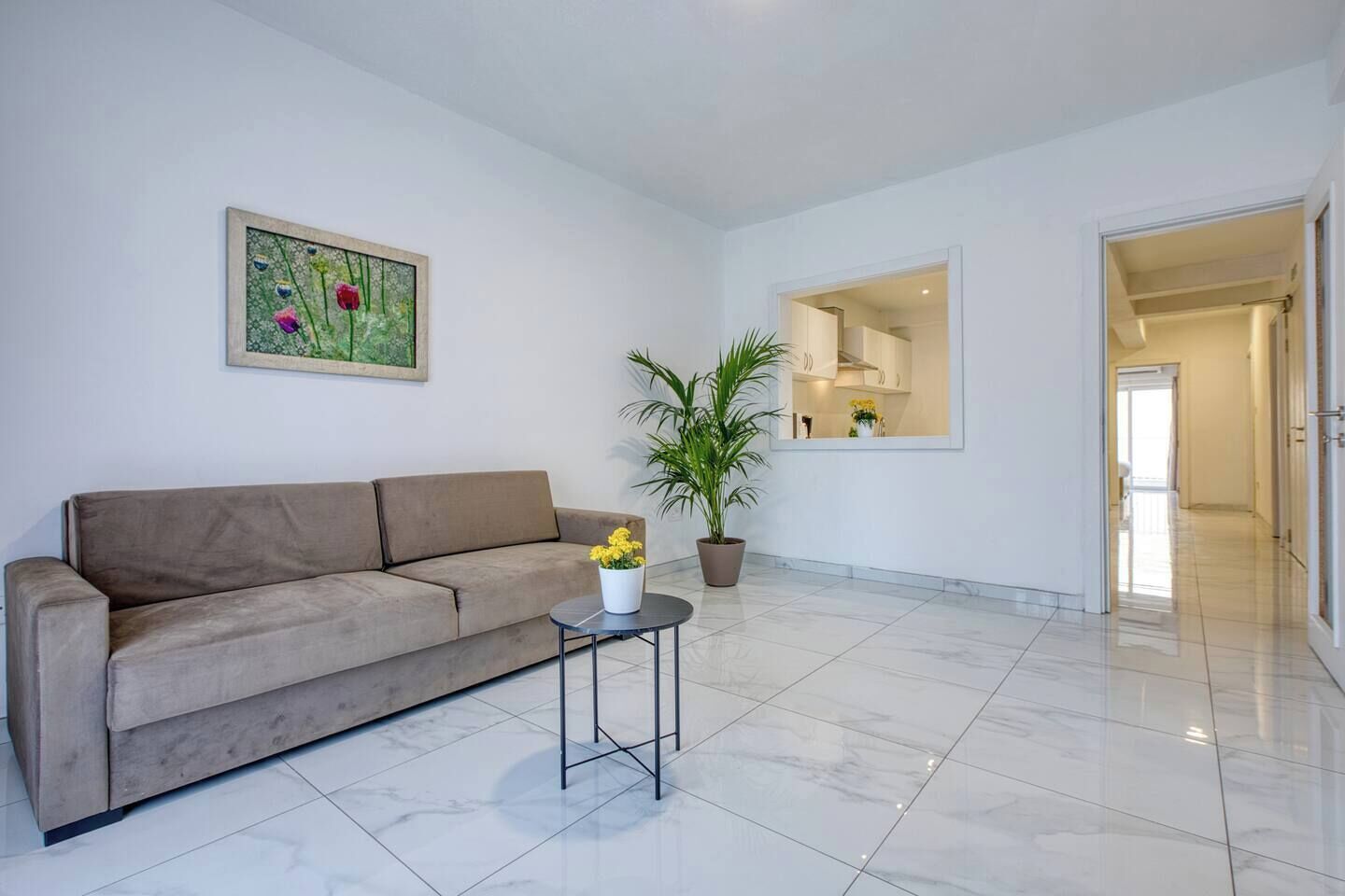 A living room with a sofa, a small table with flowers, a potted plant, and a painting on the wall.