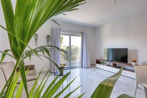 Ein modernes Wohnzimmer mit Flachbildfernseher, einer Couch, einem Esstisch und einem Balkon mit Vorhängen.