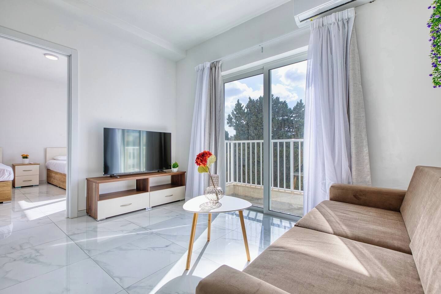 A modern living room with a flat-screen TV, a sofa, a coffee table, and a balcony with curtains.