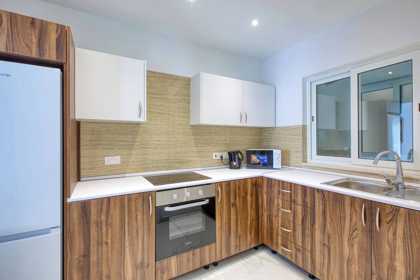 Een moderne keuken met houten kastjes, een ingebouwde oven, een magnetron en een raam boven de spoelbak.
