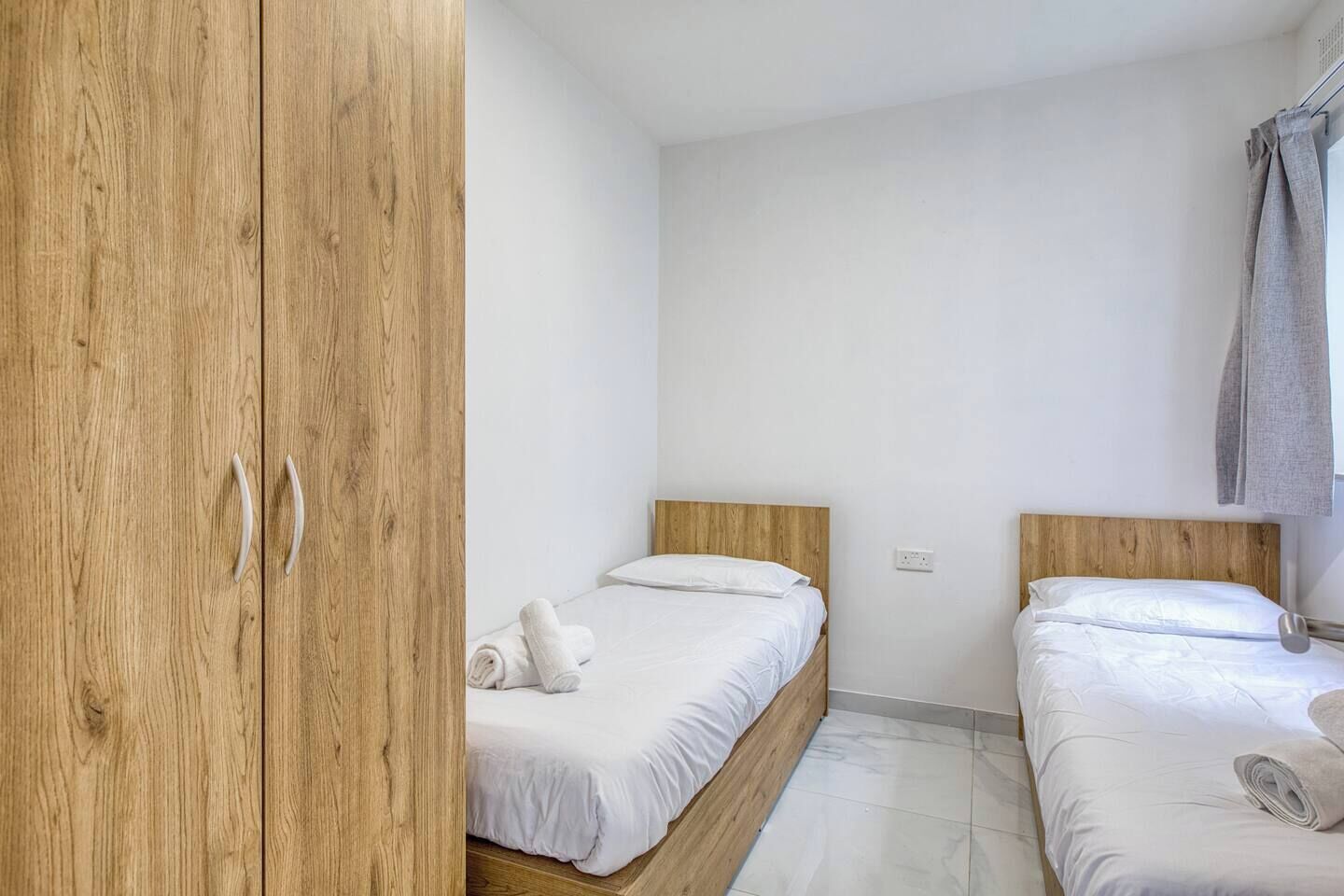 A bedroom with two single beds, wooden wardrobe, white bedding, and a grey curtain.