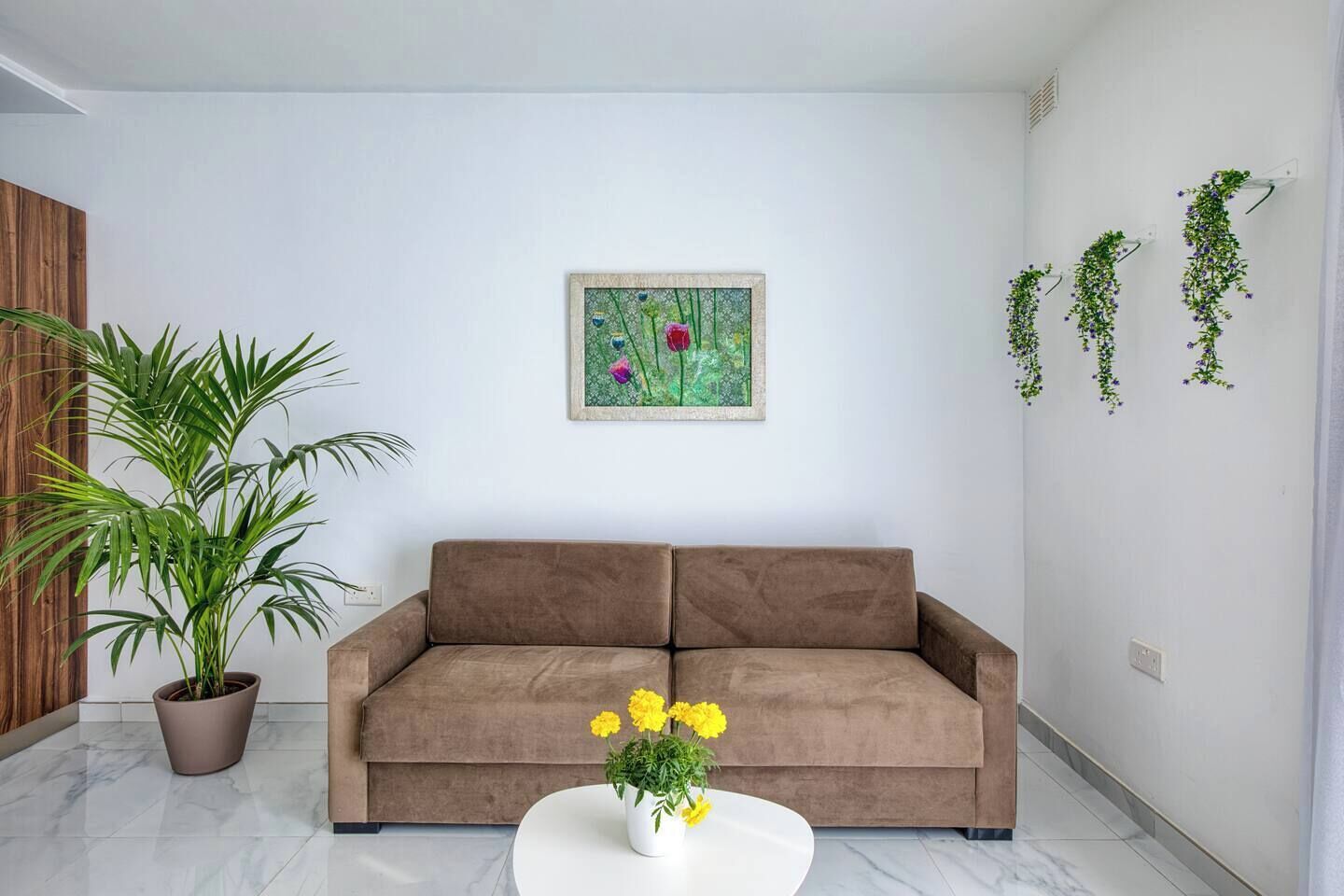 A living room with a brown sofa, a potted plant, a painting, and hanging plants on the wall.