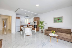Ein modernes Wohnzimmer mit Marmorboden, einem braunen Sofa, einem Esstisch mit Stühlen, einer Küche im Hintergrund und einer Zimmerpflanze.
