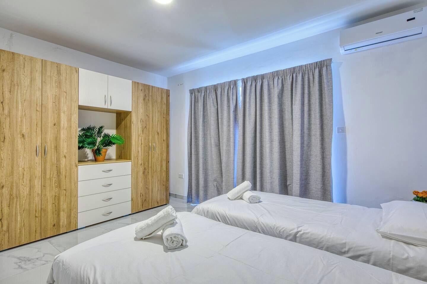 A hotel room with two beds, a wardrobe, a potted plant, and an air conditioning unit.