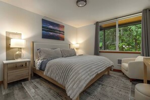 Ein modernes Schlafzimmer mit einem großen Bett, Nachttischlampen, Blick auf Bäume durch ein Fenster und einem Bild an der Wand.