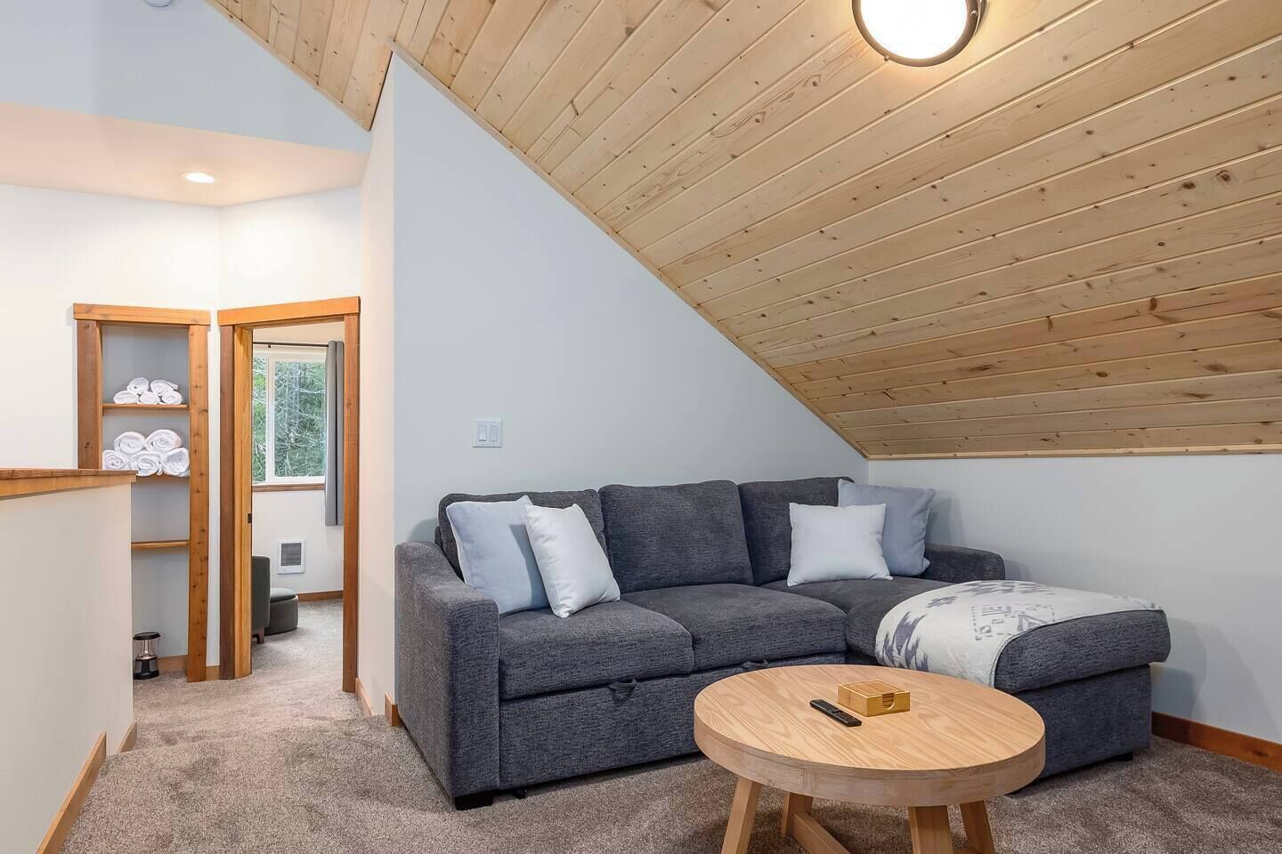 A cozy room with a grey sofa, a round wooden table, and a wooden ceiling.