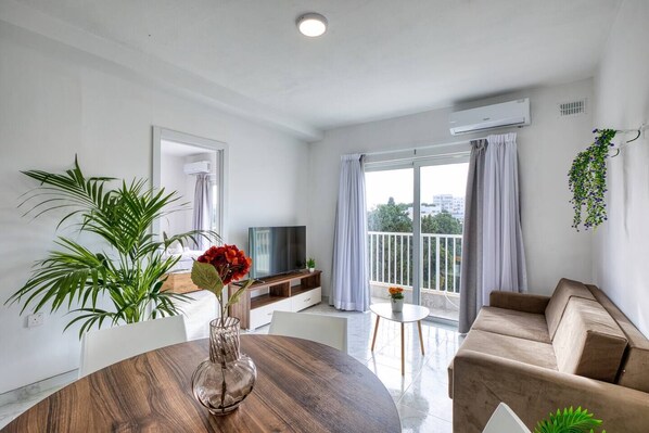 A modern living room with a round wooden table, a sofa, a TV, and a balcony with curtains. - Modern, Central 3-BR Apt with Valletta View (Tal-Pietà)