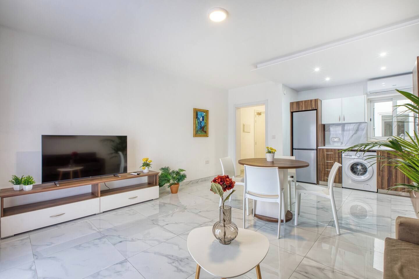 A modern living room with a flat-screen TV, a dining area with a round table, and a kitchen with a built-in washer and dryer.