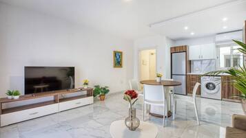 A modern living room with a flat-screen TV, a dining area with a round table, and a kitchen with a built-in washer and dryer.