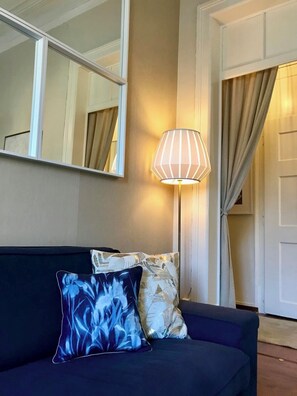 Ein dunkel gefärbtes Sofa mit gemusterten Kissen, eine Stehlampe und ein Spiegel an der Wand.