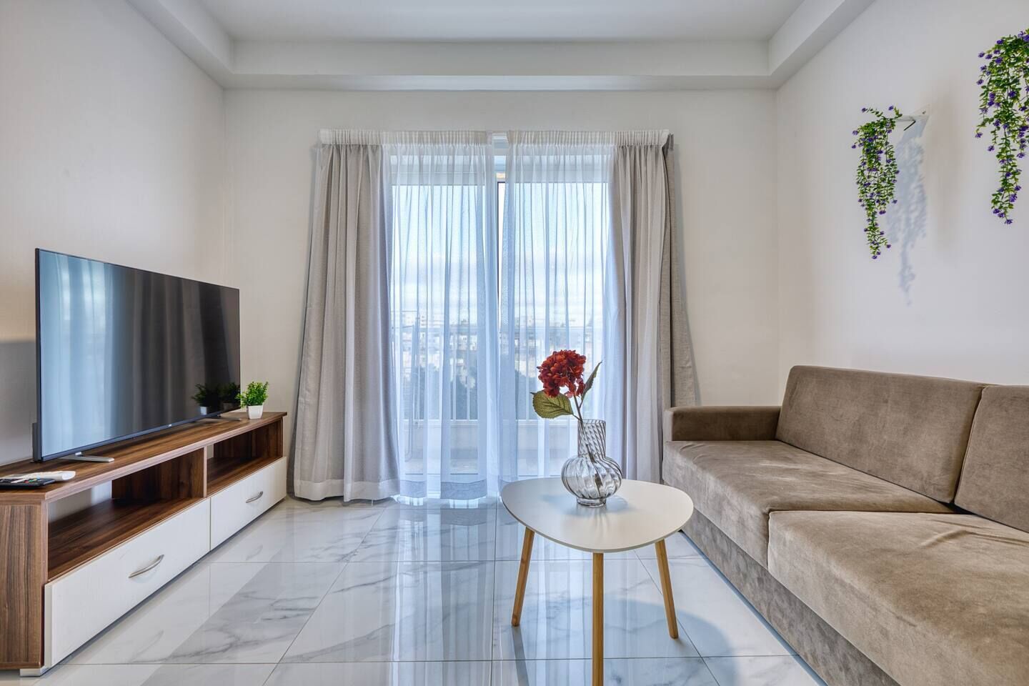 A modern living room with a flat-screen TV, a sofa, a coffee table with a vase, and sheer curtains.