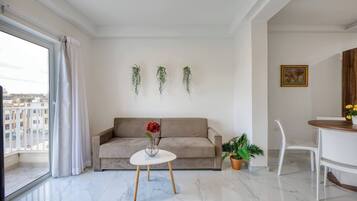 A modern living room with a sofa, a small round table, a potted plant, and a view of the city through a large window.