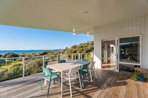 A wooden deck with a table and chairs, a glass door leading to an interior space, and a view of the ocean. - Rings Beach Retreat - Award Winning Home (Kūaotunu West)