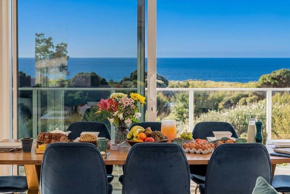 A dining table with breakfast items and a vase of flowers, overlooking a coastal view.