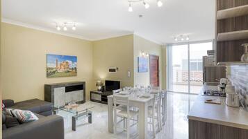 Una sala de estar moderna con un sofá, una mesa de centro de vidrio y un comedor con una mesa y sillas blancas. Hay una chimenea, un televisor de pantalla plana y un cuadro en la pared.