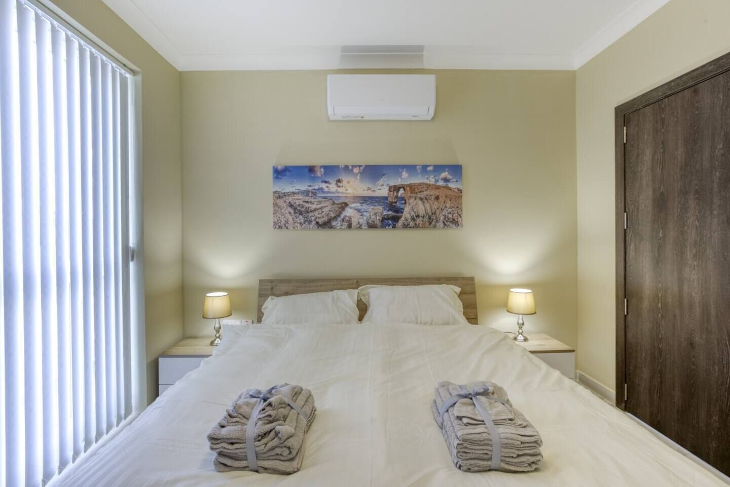 Una habitación de hotel con una cama, dos lámparas de noche, un cuadro en la pared y una puerta.