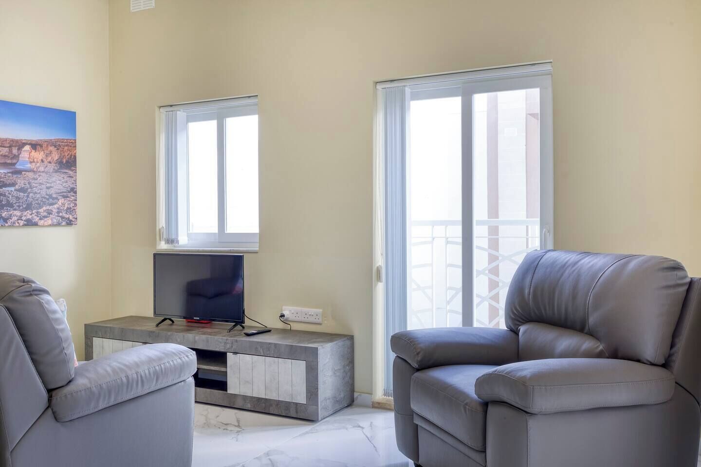 A living room with a flat-screen TV on a marble console, two grey leather armchairs, and a large window with a view of a balcony.