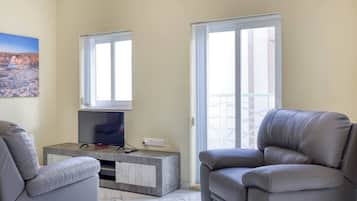 A living room with a flat-screen TV on a marble console, two grey leather armchairs, and a large window with a view of a balcony.