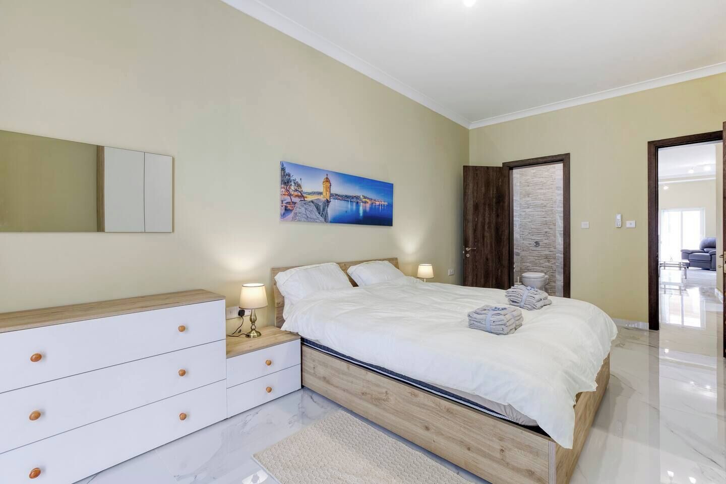 A bedroom with a bed, bedside tables, a mirror, a painting, and a door leading to another room.