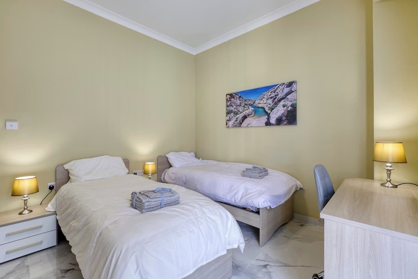 A hotel room with two beds, a desk, a chair, and a painting on the wall.