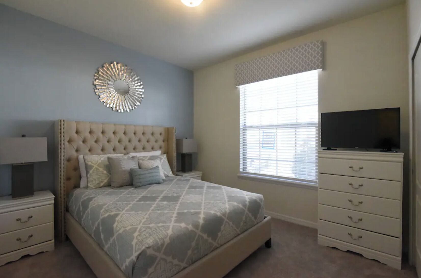 A bedroom with a tufted headboard, a bed with patterned bedding, a bedside table with a lamp, a dresser with drawers, a television on top, and a window with blinds.