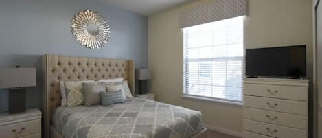 Une chambre à coucher avec une tête de lit capitonnée, un lit avec une literie à motifs, une table de chevet avec une lampe, une commode avec des tiroirs, un téléviseur posé sur le dessus, et une fenêtre avec des stores.