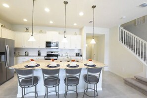 Een moderne keuken met een centraal eiland, krukken, roestvrijstalen apparatuur en hangende verlichting.