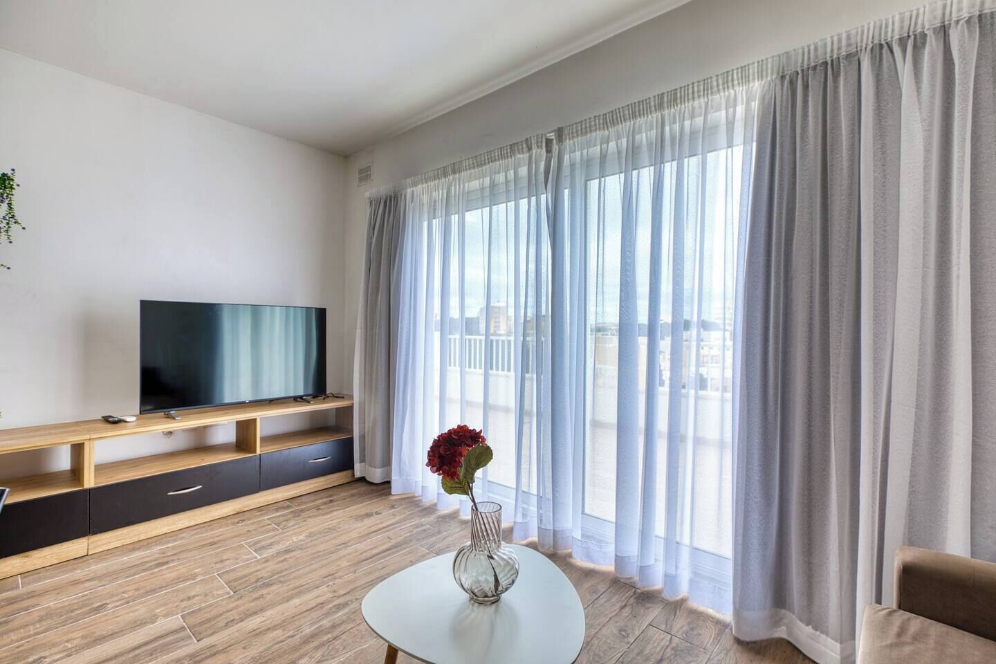 Uma sala de estar moderna com uma TV de tela plana sobre um móvel de madeira, uma mesa de centro de vidro com um vaso, e uma janela grande com cortinas translúcidas.