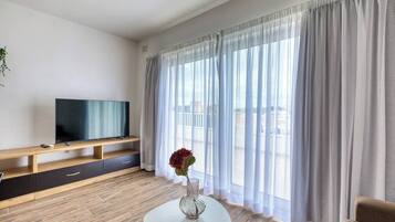Uma sala de estar moderna com uma TV de tela plana sobre um móvel de madeira, uma mesa de centro de vidro com um vaso, e uma janela grande com cortinas translúcidas.