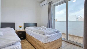 Une chambre d’hôtel dotée d’une grande porte-fenêtre coulissante donnant sur un balcon, d’un lit en bois, d’une table de chevet ornée d’un vase de fleurs et d’un climatiseur fixé au mur.