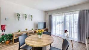 Un salon moderne avec une table ronde en bois, des chaises noires, un téléviseur à écran plat et une grande fenêtre ornée de rideaux légers.