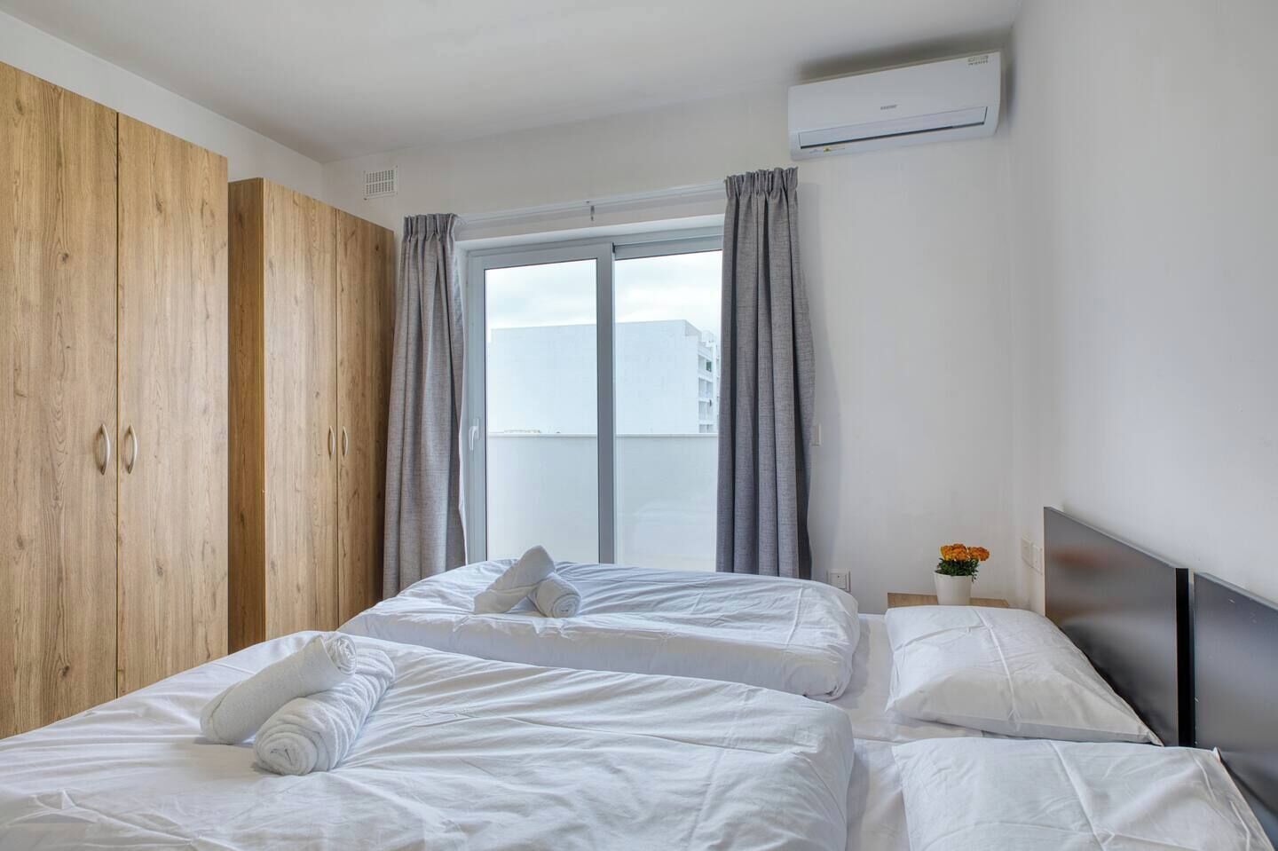 A hotel room with a large window, a wardrobe, a bed with white linens, and a small table with a vase of flowers.