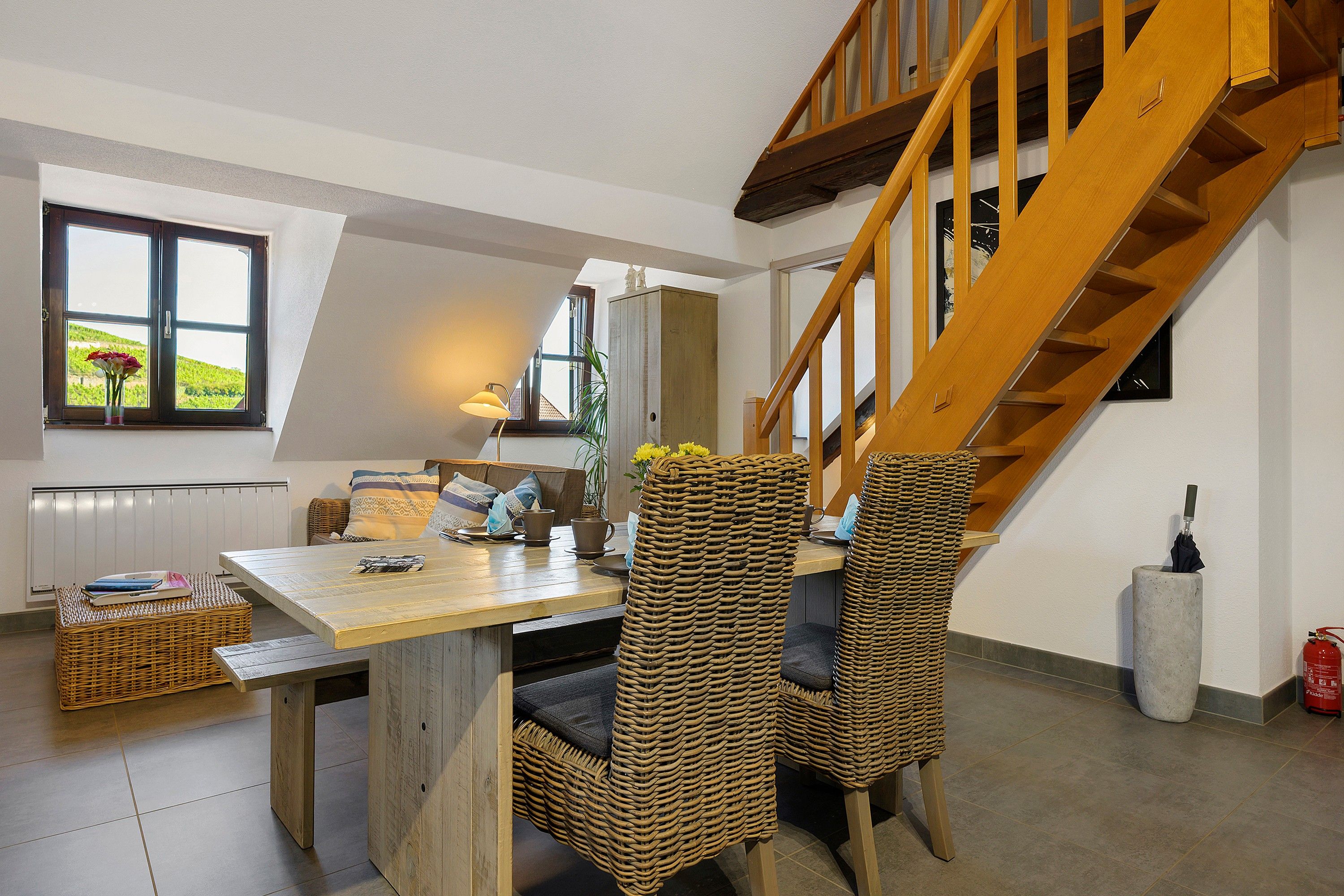Un intérieur moderne comprenant un coin repas, des chaises en osier, un escalier en bois et une vue par la fenêtre.