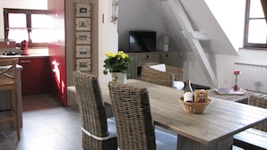 A dining area with wicker chairs and a wooden table, a kitchen with red cabinets, and a television on a cabinet. - Le Bretzel Apartment with Mountain View and Wi-Fi (Ribeauvillé)