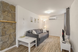 A modern living room with a stone wall, a grey sofa, a white table, and a mounted TV. - 2 beds | AC | Smart TV Netflix | Près Vieux-Québec (Québec)