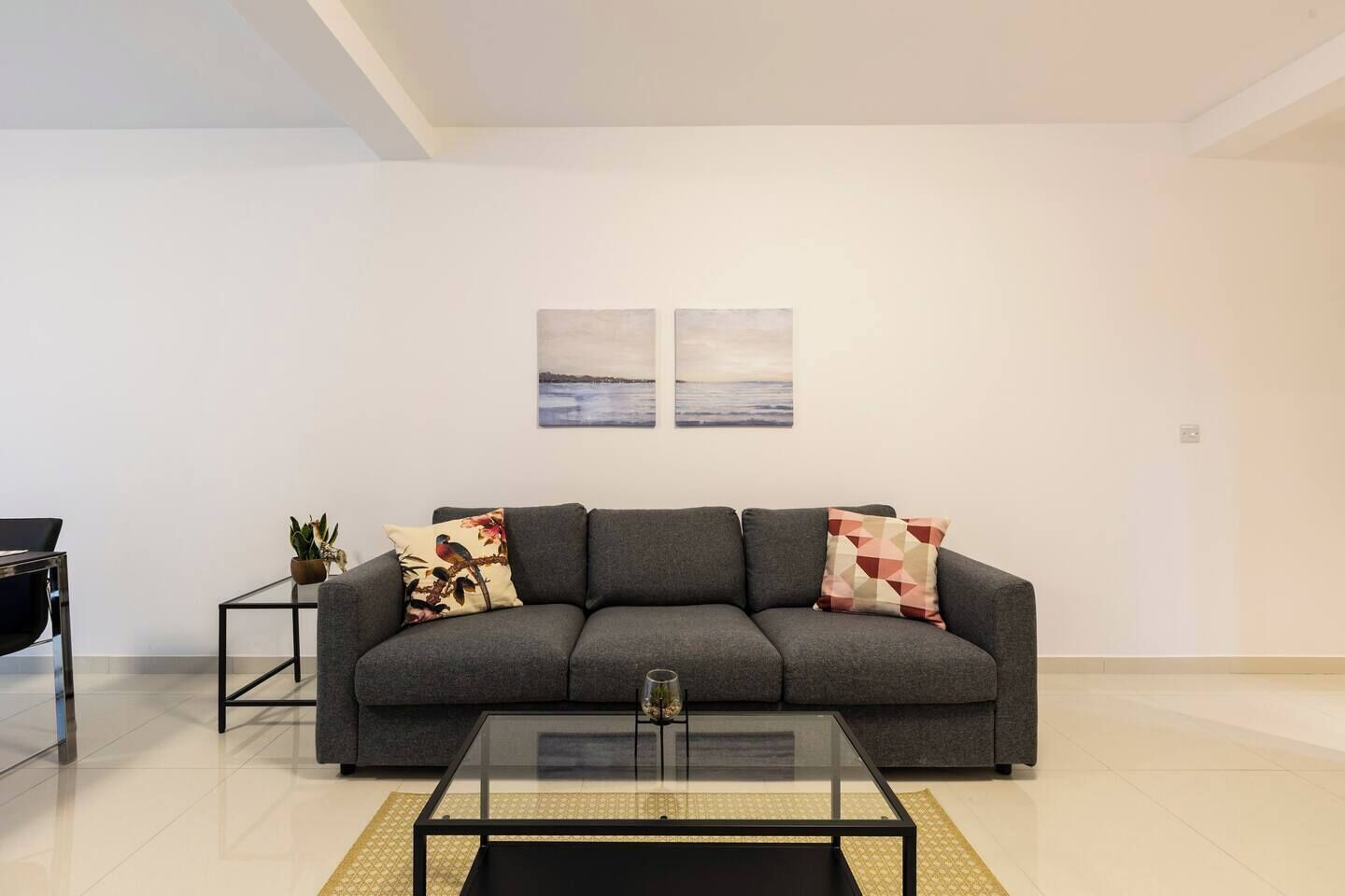 Una sala moderna con un sofá gris, dos cojines, una mesa de centro de vidrio y una mesita auxiliar con una planta.