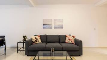 Una sala moderna con un sofá gris, dos cojines, una mesa de centro de vidrio y una mesita auxiliar con una planta.
