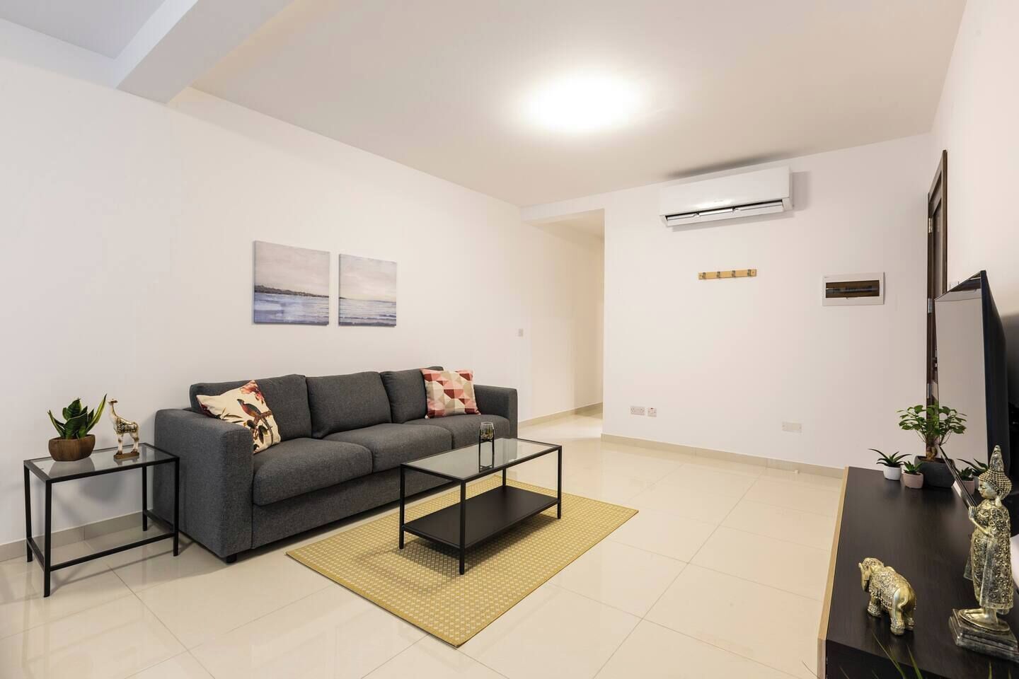 Una sala moderna con un sofá gris, una mesa de centro de vidrio y una mesita auxiliar con una planta.