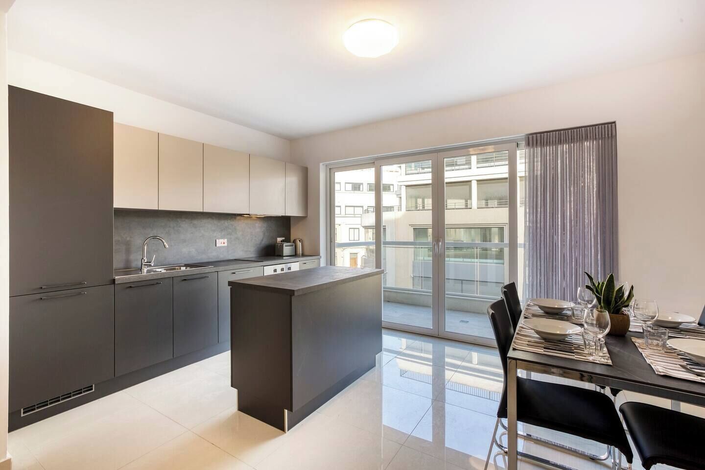 Una cucina moderna con isola centrale, zona pranzo con tavolo apparecchiato per due e un'ampia porta scorrevole in vetro che dà su un balcone.