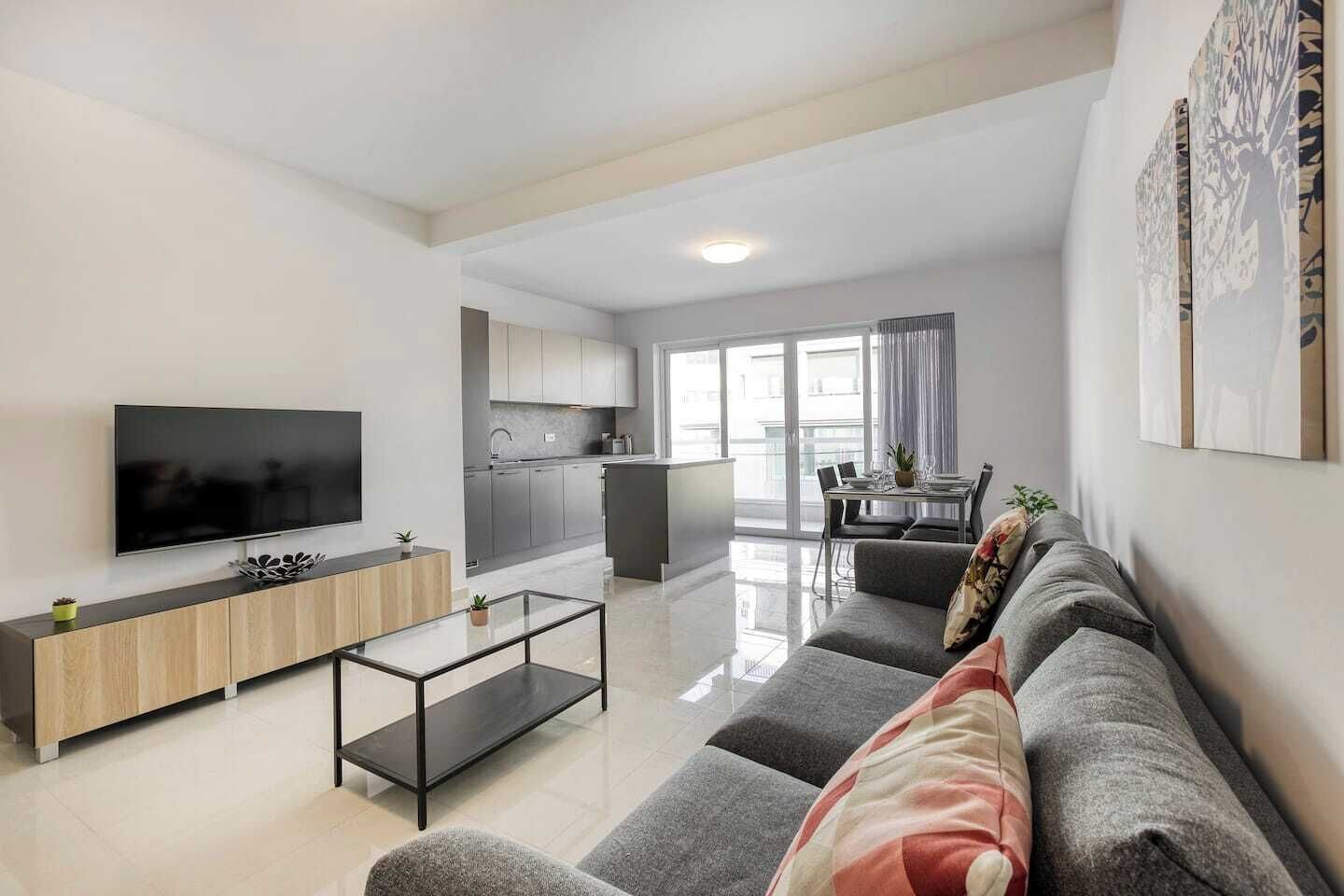 Un soggiorno moderno con televisione a schermo piatto, un tavolino in vetro e un divano grigio con cuscini decorativi.
