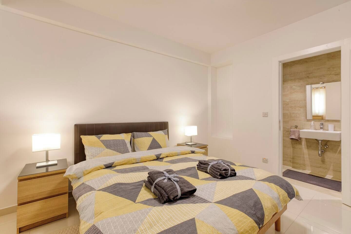 Una habitación de hotel con cama, mesitas de noche, lámpara y baño con lavabo y espejo.