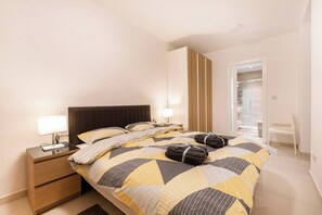 Ein Hotelzimmer mit Bett, Nachttischen, einer Lampe, einem Kleiderschrank und einem durch eine Glastür einsehbaren Badezimmer.