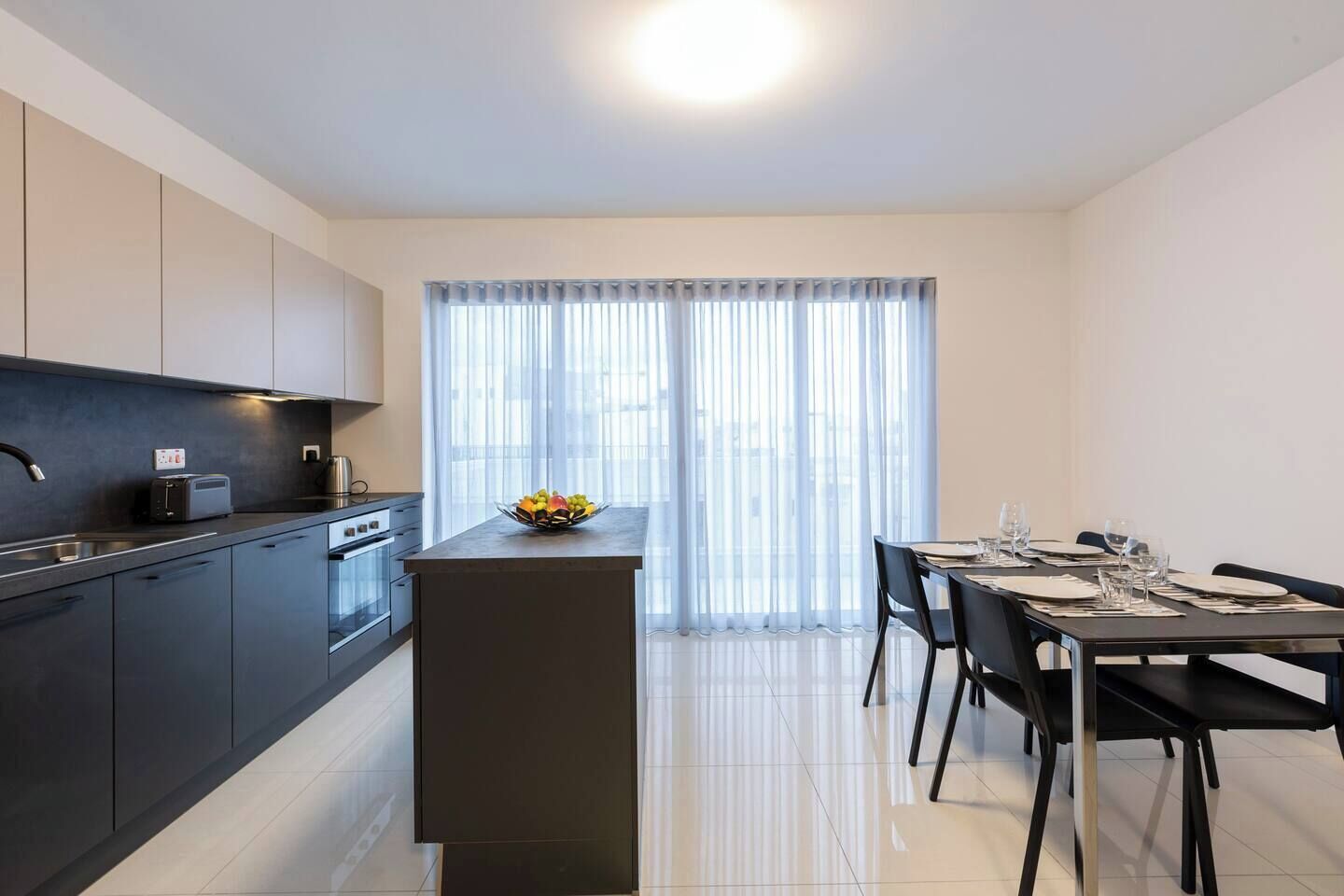 Una cucina moderna con isola centrale, zona pranzo con tavolo apparecchiato per quattro e un'ampia finestra con tende leggere.