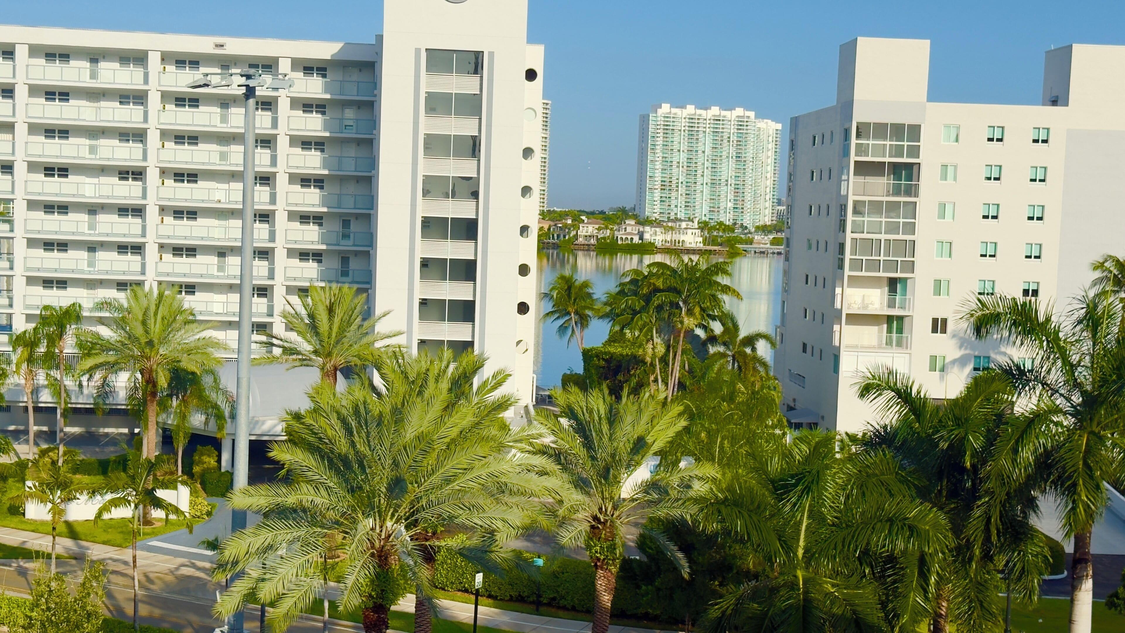 A modern high-rise building with multiple balconies, surrounded by palm trees and a body of water.