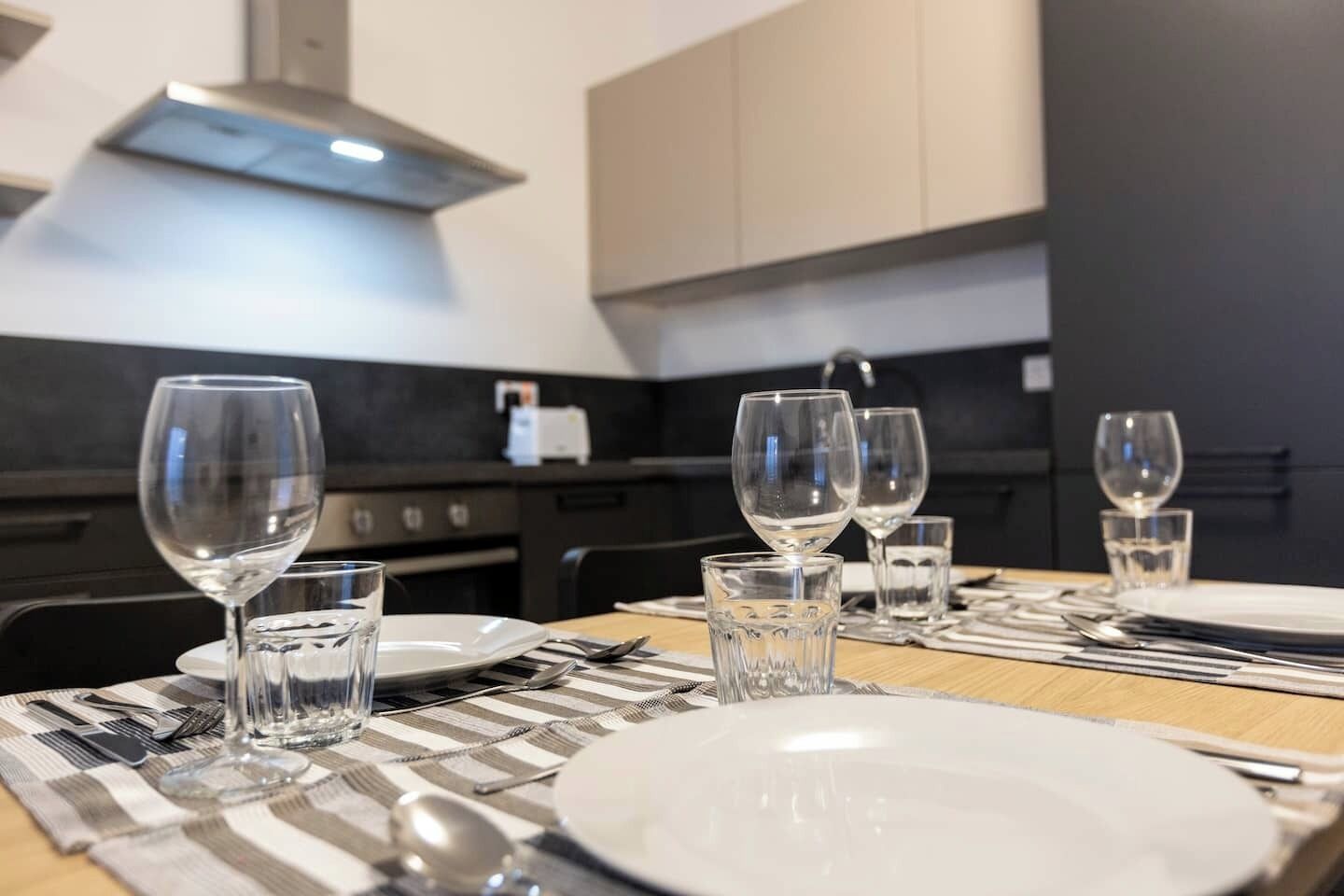 Una mesa de comedor dispuesta con platos, vasos y cubiertos, con una cocina moderna al fondo.