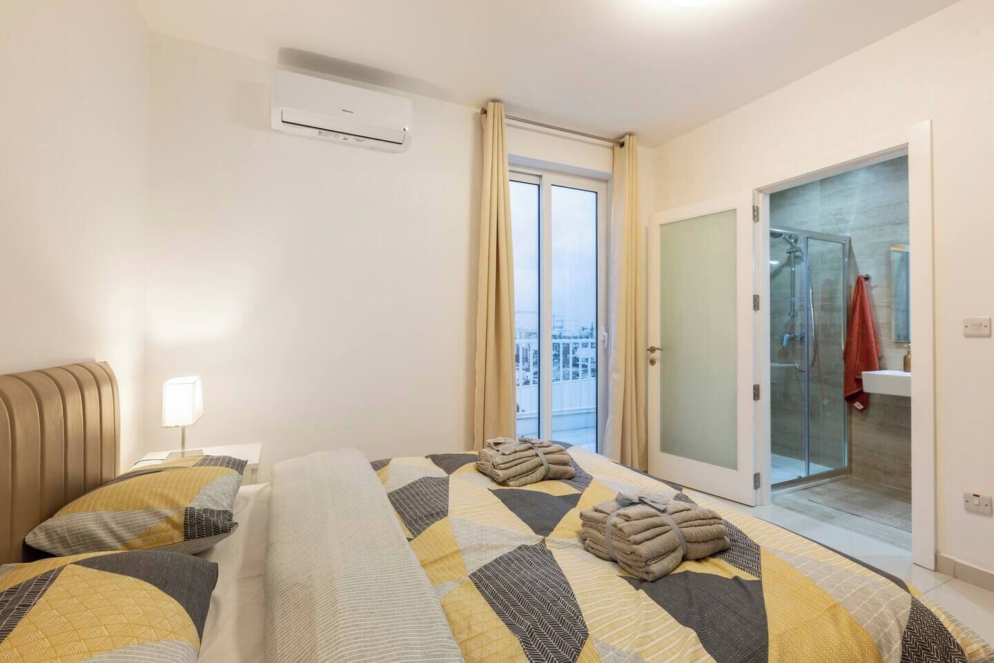 Camera d'albergo con un letto, una lampada da comodino, un condizionatore d'aria, un bagno con porta in vetro e vista sulla città.