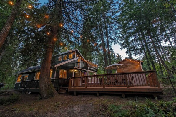 A cabin with a deck and string lights, surrounded by tall trees. - Harry's Hideaway | Hot Tub | Fire pit | Mt Rainier (Packwood)