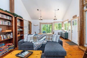 Ein gemütliches Wohnzimmer mit einem grauen Ecksofa, einem Holztisch und einem Bücherregal voller Bücher. Zwei Pendelleuchten hängen von der Decke.