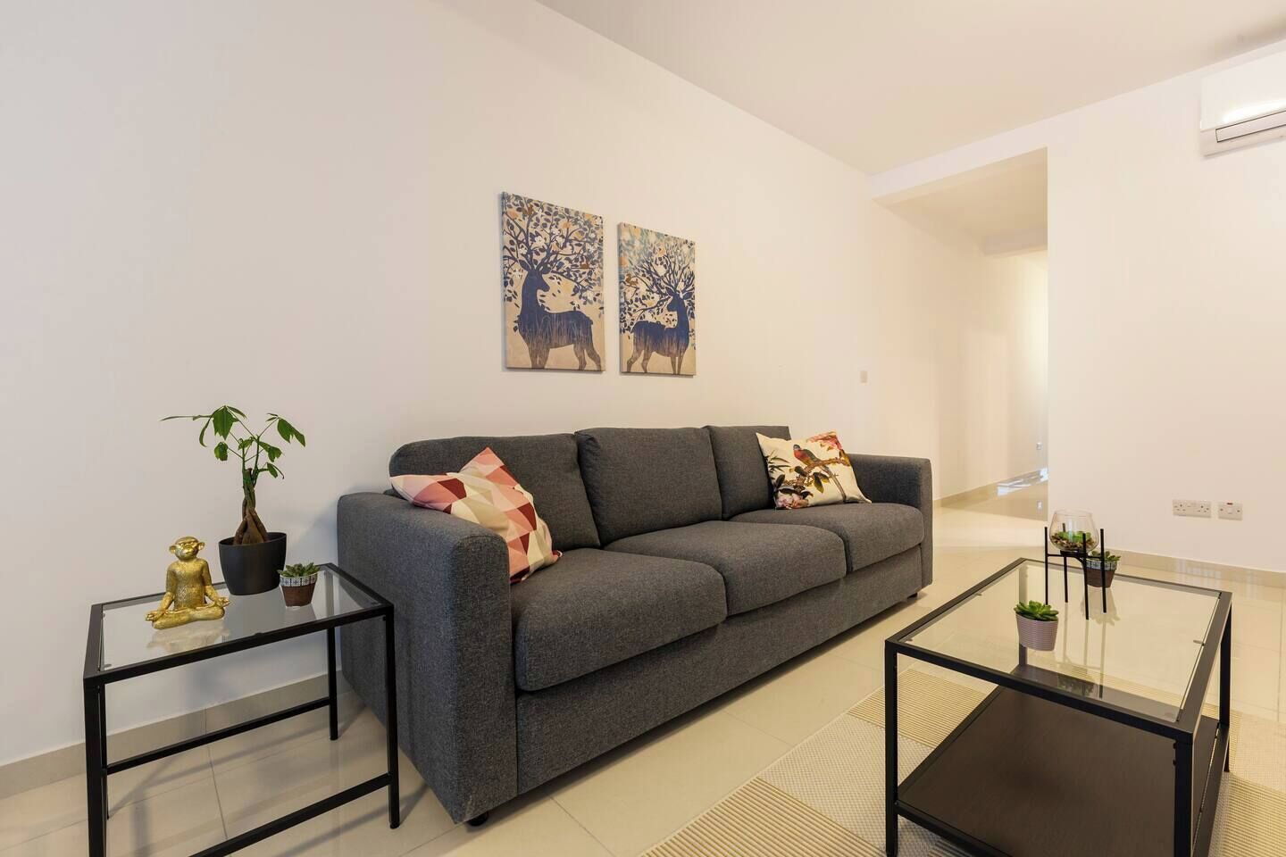 Un salon moderne avec un canapé gris, deux tables d’appoint, une table basse en verre et des objets décoratifs, notamment une statue de Bouddha et une plante en pot.