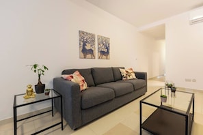 A modern living room with a grey sofa, two side tables, a glass coffee table, and decorative items including a Buddha statue and a potted plant.